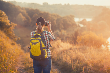tourist girl with a backpack and a camera walks through a picturesque meadow and photographs the picturesque landscape. Tourism, outdoor activities - obrazy, fototapety, plakaty
