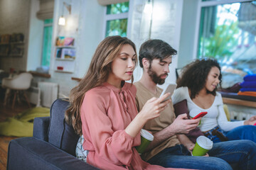 Two girls and guy sitting on sofa with smartphones and coffee.