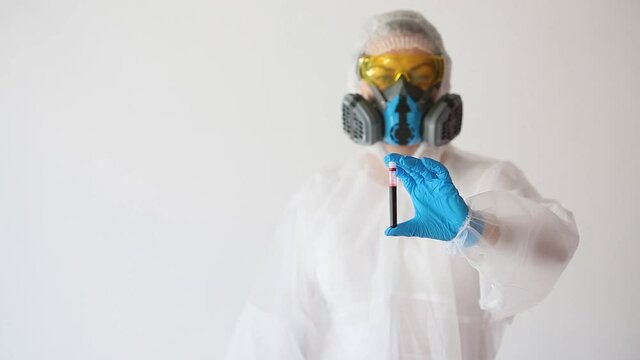 Doctor in a white protective suit and respirator shows a blood sample in a test tube and a teath sign. Focus on the hands