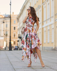 Young beautiful elegant tall slim woman with natural makeup and wavy brunete hair wearing colorful dress and sunglassses walking in the city on a summer day and holding handbag
