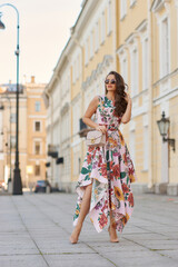 Fototapeta na wymiar Young beautiful elegant tall slim woman with natural makeup and wavy brunete hair wearing colorful dress and sunglassses walking in the city on a summer day and holding handbag