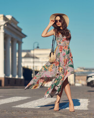 Young beautiful elegant tall slim woman with natural makeup and wavy brunete hair wearing colorful dress, salty hat and sunglassses walking in the city on a summer day and holding handbag