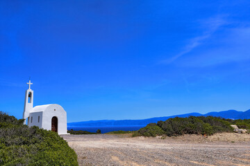 White Greek Orthodox chapel or church and road to it on hilltop of seashore against clear blue sky on sunny day.