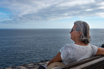 Fototapeta premium Senior lady with gray hair sitting on a bench in front to the ocean - relaxed people in sea excursion, freedom and retirement concept