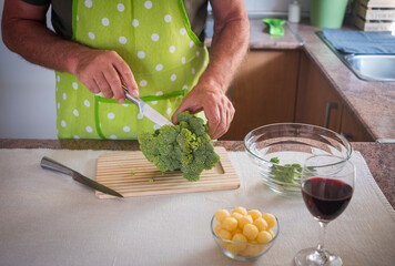 A senior man cleans and cuts broccoli in the kitchen, a snack and a glass of red wine near him - healthy, vegan and vegetarian lifestyle concept