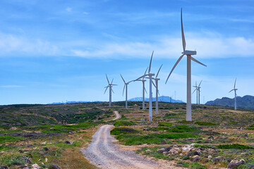 Windmill turbine farm on a hilltop in colorful landscape with winding road against blue sky on clear sunny summer day.