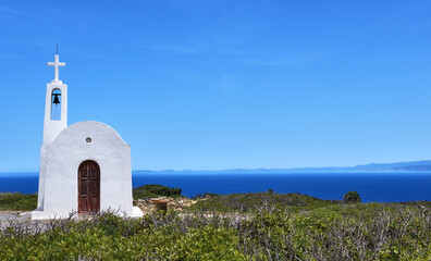 White Greek Orthodox chapel or church on hilltop of seashore against clear blue sky on sunny day.