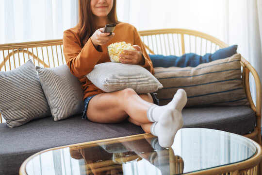 Closeup image of a beautiful young woman eating popcorn and searching channel with remote control to watch tv while sitting on sofa at home