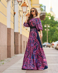 Elegant brunette woman with long wavy hair wearing long purple sundress, standing and posing at city street on a summer day. Full length portrait