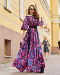 Elegant brunette woman with long wavy hair wearing long purple sundress, standing and posing at city street on a summer day. Full length portrait