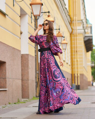 Naklejka premium Elegant brunette woman with long wavy hair wearing long purple sundress, standing and posing at city street on a summer day. Full length portrait