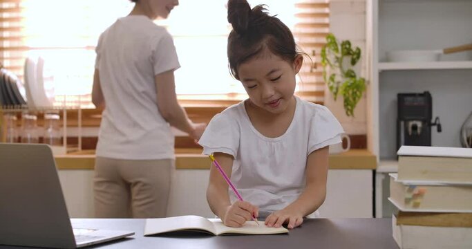 Cute concentrated little kid girl studying online in the kitchen at home. Young asian primary school girl studying at table, doing homework. Children remote education on quarantine concept.
