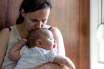 Photo of little baby girl sleeping in her mother's arms. Women wearing casual clothes carrying and kissing her baby daughter in the kitchen. The baby is sleeping and resting on her mother's shoulder