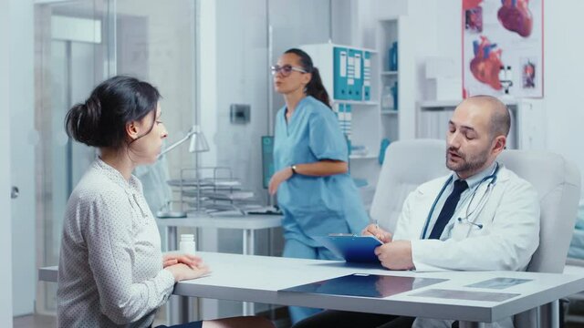 Doctor questioning young female patient in his office, writing answears in clipboard. Healthcare in modern hospital or private clinic, disease prevention and consultation in medic office treatment