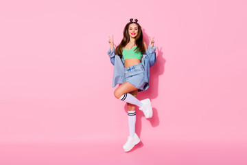 Full length photo of pretty funky lady jumping high up showing v-sign symbols wear denim jacket green crop top naked belly skirt knee socks shoes isolated pastel pink color background