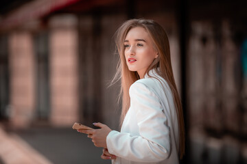 Portrait of Beautiful young business woman with phone at the hand on the summer street. Female model with long hair wearing at white suit standing at the city