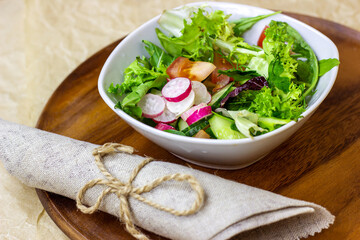 Bright fresh green salad leaves with red tomatoes, cucmbers and radish in the white bowl on wooden background.