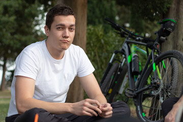 Young man resting sitting next to his bike in public, green grass and trees around him. Bicycle rider enjoying a warm day