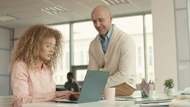 Young woman with curly hair sitting at office sek working on laptop, her male colleague sitting next to her and listening to her