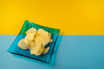 Closeup shot of plain potato chips in a blue glass plate on a yellow blue background