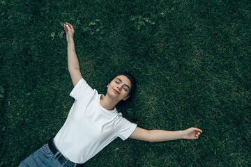 Caucasian girl 20-25 years old lies on the green grass on a summer day smiling, arms outstretched,...