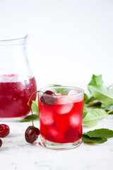 Cold summer drink with red ice in a glass glass on a light background. Cherry berries and green leaves in the background.