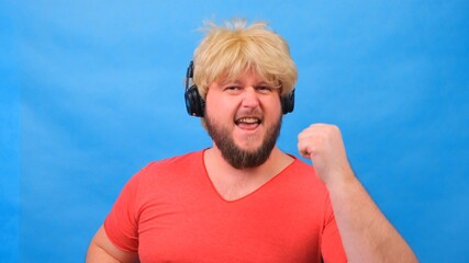 Naklejka premium freaky fat man in a wig and a pink T-shirt in his hands listens to music on headphones and dances on a blue background