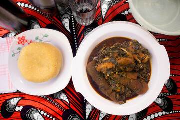 A delicious meal of Nigerian vegetable soup cooked with assorted meat, dried fish, cow skin (ponmo or kpomo or pomo) and Garri or Eba. Served on a colorful red and white African pattern table cloth