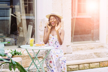 Portrait of a relaxed young african american woman drinking lemonade, eating dessert, sitting at cozy outdoor cafe with green plant talking on cell phone.. Sun glare effect.