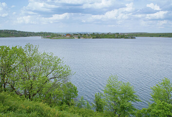 Landscape on green shore in May. Blue cloudy sky over horizon. Green trees against blue water. View from the high bank.