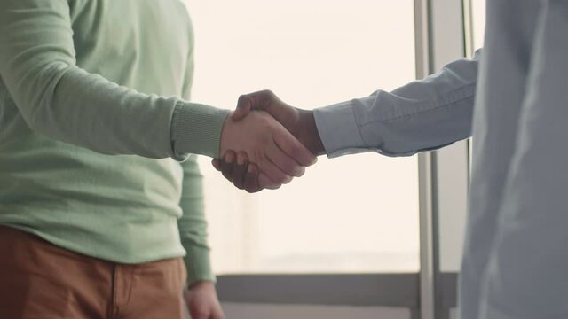 Two unrecognizable men working in modern office greeting each other with handshake, side view medium close-up shot