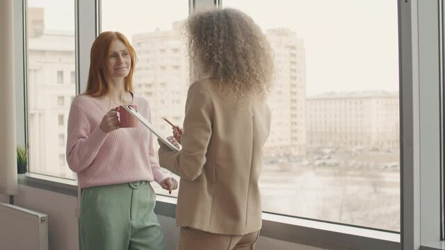 Two stylish businesswomen of different age standing together at office window talking about something funny and smiling during coffee break