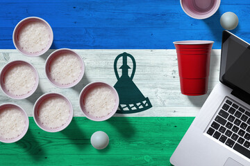 Lesotho flag concept with plastic beer pong cups and laptop on national wooden table, top view. Beer Pong game.