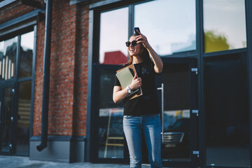 Happy cheerful female student with modern smartphone device in hand feeling excited on urban setting,successful woman writer with textbooks waiting friend outdoors raising hands for greeting or waving