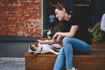 Obraz premium Positive hipster girl in casual wear looking at yawn purebred french bulldog lying on city bench and feeling happy from spending time together, smiling caucasian woman enjoying leisure with lovely dog