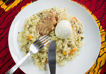 A tasty white plate of Nigerian fried rice containing sliced carrots, green beans and peppers with a piece of turkey meat and boiled egg on top. A fork and knife is placed on the side