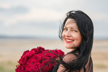 Portrait of a happy girl with long black hair with a large bouquet of red roses on nature.
