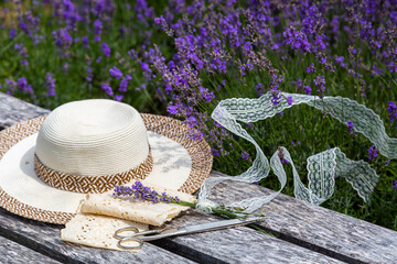 A straw hat, lice ribbons, vintage scissors and little lavender bouquet on wooden bench near lavender bushes.