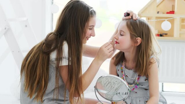 Pretty teenage girl painting lips of cute little sister indoors