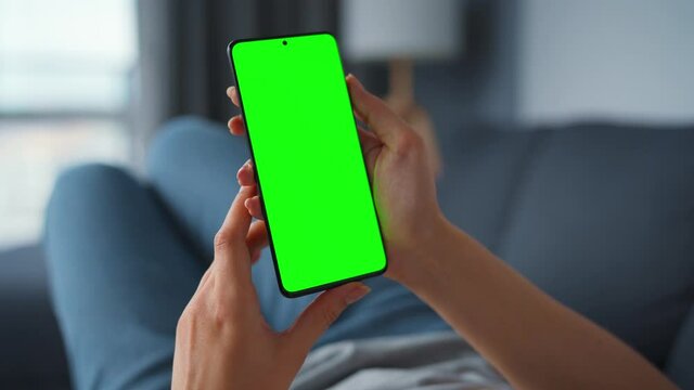 Woman at home lying on a sofa and using smartphone with green mock-up screen in vertical mode. Girl browsing Internet, watching content, videos, blogs. POV.