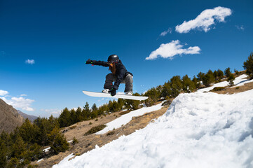 Naklejka na ściany i meble Stylish young girl snowboarder does the trick in jumping from a snow kicker against the blue sky clouds and mountains in the spring.