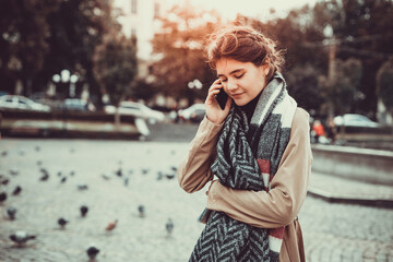 Street style portrait of smiling beautiful girl in autumn outfit, talking on phone, enjoying sunny day.