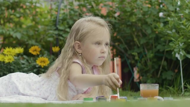 Blond girl draws paints on the nature. Girl 5 years old lies on the grass and paints a picture
