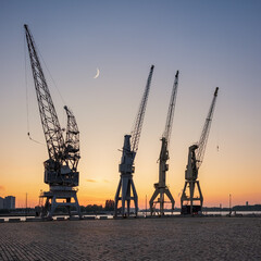 Obraz premium Old harbor crane silhouettes under a crescent moon. The cranes are part of the collection of the MAS museum.