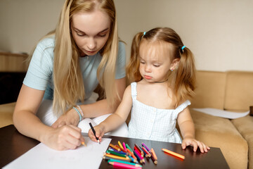 Young beautiful mother blonde and her daughter 2 years old draw with pencils on the sofa in the living room at home