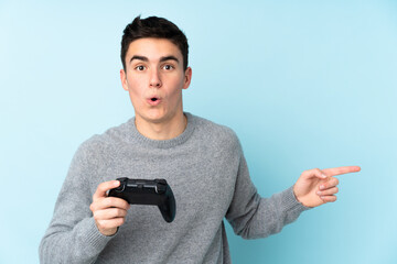 Teenager caucasian man playing with a video game controller isolated on blue background surprised and pointing side