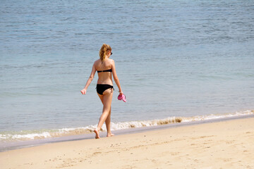 Fototapeta na wymiar Barefoot girl in bikini walking by the sand with sandals and smartphone in hand. Beach vacation on tropical sandy coast, slim female body, summer leisure