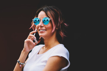 Closeup image of cute smiling hipster girl in stylish sunglasses communicating with friends via smartphone application.Happy female person talking on modern telephone during summer walk outside