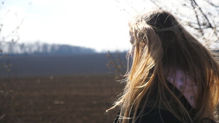 Small smiling girl with blowing blonde hair walks on the road near plowed field. Happy kid with...