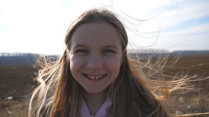 Close up of happy small girl looks into camera and straightens her blowing blonde hair against the background of plowed field. Portrait of joyful female kid stands in the ploughed meadow and laughs